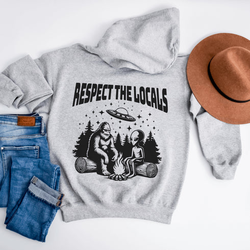 Our Respect the Locals Hoodie features the ultimate forest hangout: Bigfoot and an alien, just two mysterious dudes chilling on logs around a crackling campfire.