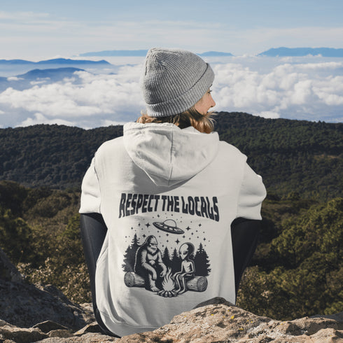 Our Respect the Locals Hoodie features the ultimate forest hangout: Bigfoot and an alien, just two mysterious dudes chilling on logs around a crackling campfire.