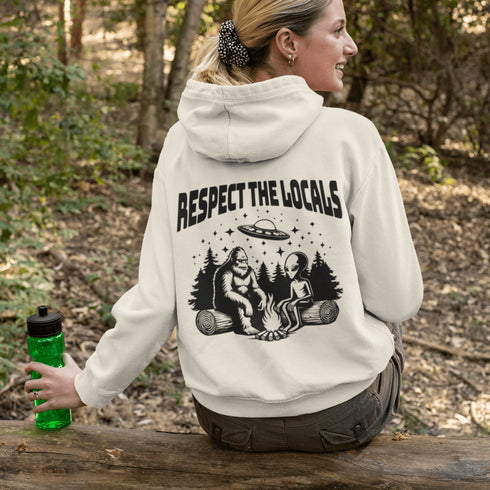 Our Respect the Locals Hoodie features the ultimate forest hangout: Bigfoot and an alien, just two mysterious dudes chilling on logs around a crackling campfire.