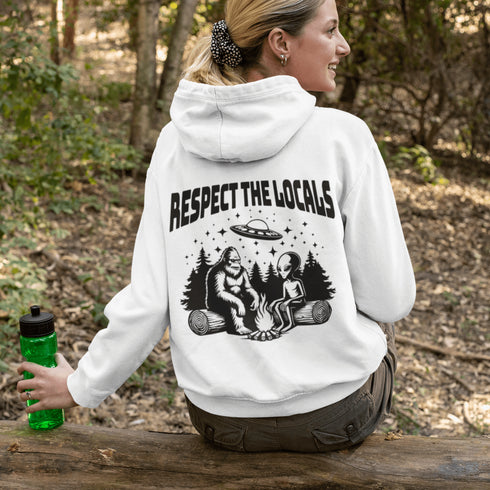 Our Respect the Locals Hoodie features the ultimate forest hangout: Bigfoot and an alien, just two mysterious dudes chilling on logs around a crackling campfire.