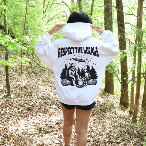 Our Respect the Locals Hoodie features the ultimate forest hangout: Bigfoot and an alien, just two mysterious dudes chilling on logs around a crackling campfire.