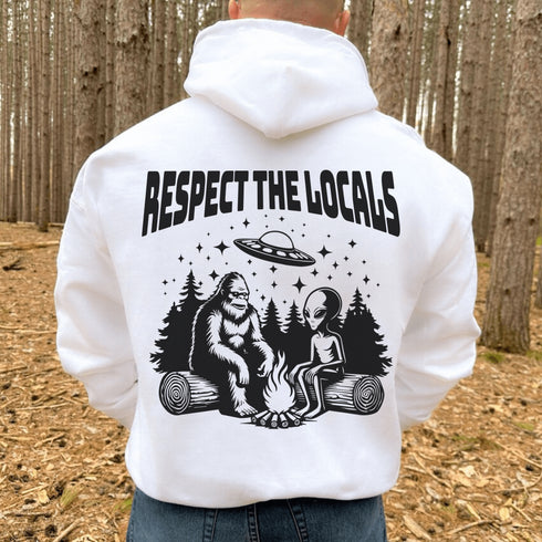 Our Respect the Locals Hoodie features the ultimate forest hangout: Bigfoot and an alien, just two mysterious dudes chilling on logs around a crackling campfire.