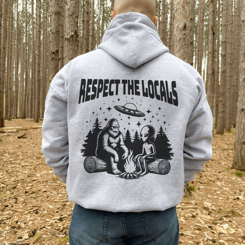 Our Respect the Locals Hoodie features the ultimate forest hangout: Bigfoot and an alien, just two mysterious dudes chilling on logs around a crackling campfire.