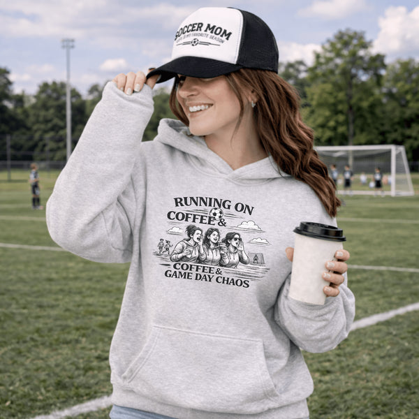 Soccer mom hoodie with “Running on Coffee and Game Day Chaos” design featuring moms cheering on the sidelines with kids playing soccer in the background