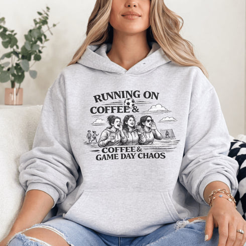 Soccer mom hoodie with “Running on Coffee and Game Day Chaos” design featuring moms cheering on the sidelines with kids playing soccer in the background