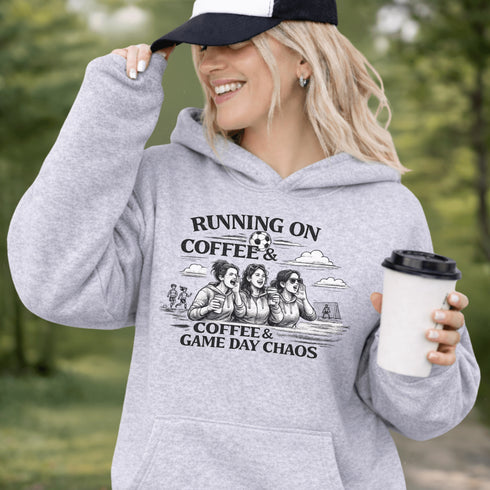 Soccer mom hoodie with “Running on Coffee and Game Day Chaos” design featuring moms cheering on the sidelines with kids playing soccer in the background
