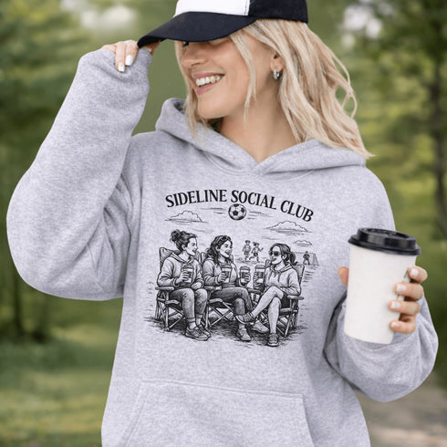 Soccer mom hoodie with “Sideline Social Club” design featuring moms sitting in lawn chairs drinking coffee while watching kids play soccer in the background