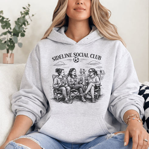 Soccer mom hoodie with “Sideline Social Club” design featuring moms sitting in lawn chairs drinking coffee while watching kids play soccer in the background
