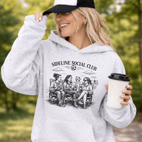 Soccer mom hoodie with “Sideline Social Club” design featuring moms sitting in lawn chairs drinking coffee while watching kids play soccer in the background