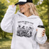 Soccer mom hoodie with “Sideline Social Club” design featuring moms sitting in lawn chairs drinking coffee while watching kids play soccer in the background