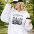 Soccer mom hoodie with “Sideline Social Club” design featuring moms sitting in lawn chairs drinking coffee while watching kids play soccer in the background