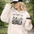 Soccer mom hoodie with “Sideline Social Club” design featuring moms sitting in lawn chairs drinking coffee while watching kids play soccer in the background