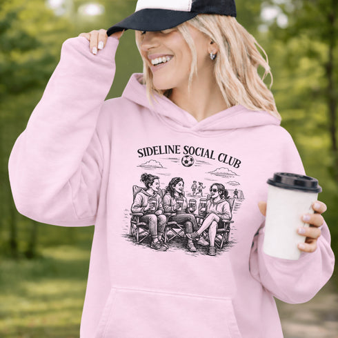 Soccer mom hoodie with “Sideline Social Club” design featuring moms sitting in lawn chairs drinking coffee while watching kids play soccer in the background