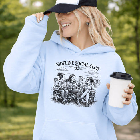Soccer mom hoodie with “Sideline Social Club” design featuring moms sitting in lawn chairs drinking coffee while watching kids play soccer in the background