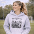 Soccer mom hoodie with “Sideline Social Club” design featuring moms sitting in lawn chairs drinking coffee while watching kids play soccer in the background