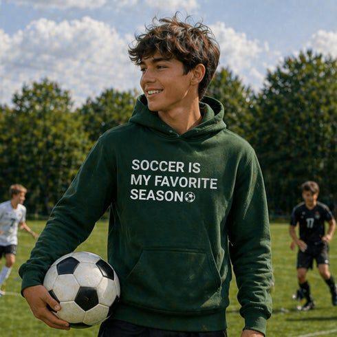 Person wearing “Soccer Is My Favorite Season” hoodie holding a soccer ball on a soccer field during a game, casual soccer apparel