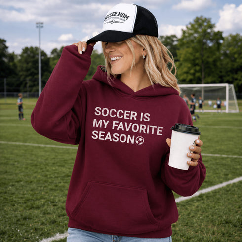 Person wearing a maroon “Soccer Is My Favorite Season” hoodie holding a coffee cup on a soccer field during a game, casual soccer apparel