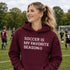 Woman wearing a Maroon “Soccer Is My Favorite Season” hoodie on a soccer field during a game, casual soccer apparel