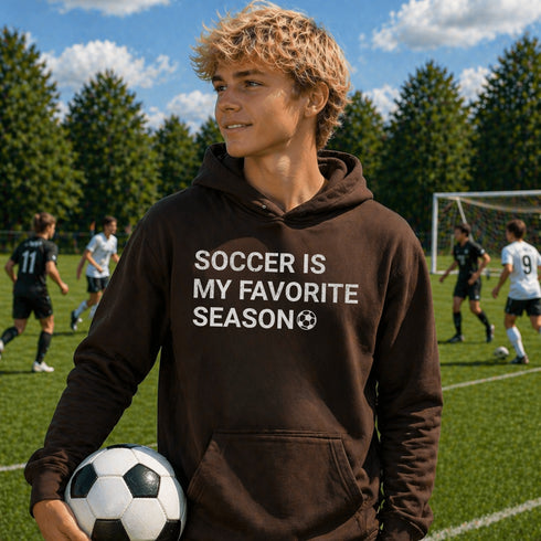 Person wearing “Soccer Is My Favorite Season” hoodie holding a soccer ball on a soccer field during a game, casual soccer apparel