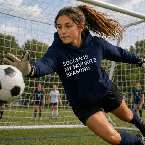 Female Soccer goalie wearing a Navy “Soccer Is My Favorite Season” hoodie stopping a soccer ball from going into the net during a game, casual soccer apparel