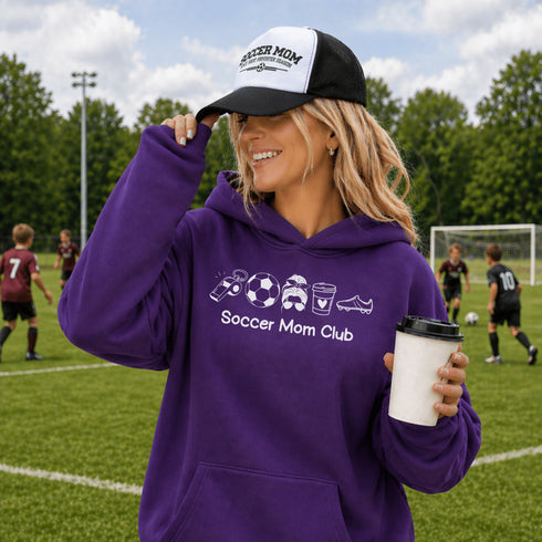 Woman wearing a purple 'Soccer Mom Club' hoodie and black and white cap on a soccer field.