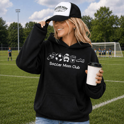 Woman wearing a black hoodie with 'Soccer Mom Club' text and holding a coffee cup on a soccer field.