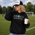 Woman wearing a black hoodie with 'Soccer Mom Club' text and holding a coffee cup on a soccer field.