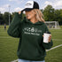 Woman wearing a green 'Soccer Mom Club' hoodie and cap, holding a coffee cup on a soccer field.