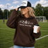 Woman wearing a 'Soccer Mom Club' hoodie and cap on a soccer field.