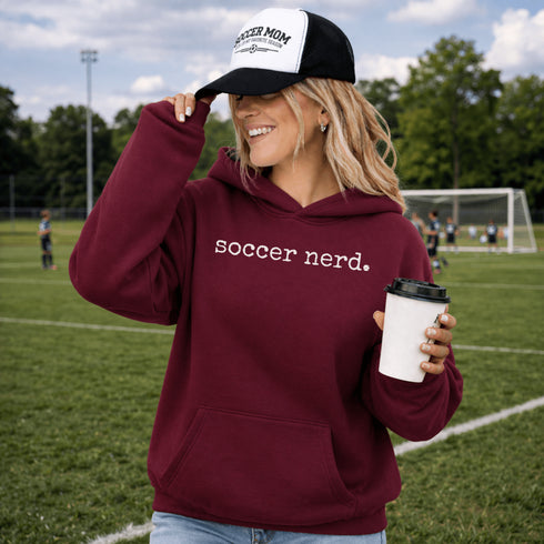 Person wearing “Soccer Nerd” hoodie holding a soccer ball on a soccer field during a game, casual athletic soccer apparel