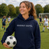 Person wearing “Soccer Nerd” hoodie holding a soccer ball on a soccer field during a game, casual athletic soccer apparel