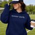 Person wearing “Soccer Nerd” hoodie holding a soccer ball on a soccer field during a game, casual athletic soccer apparel