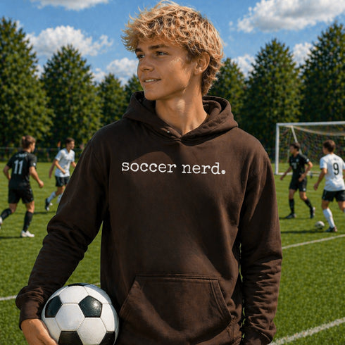 Person wearing “Soccer Nerd” hoodie holding a soccer ball on a soccer field during a game, casual athletic soccer apparel