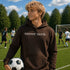 Person wearing “Soccer Nerd” hoodie holding a soccer ball on a soccer field during a game, casual athletic soccer apparel
