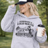 woman wearing grey hoodie with “Somebody’s Loud Mouth Soccer Mama” design featuring mom yelling on soccer sideline with kids playing in background