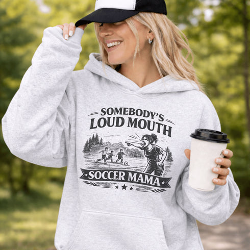 woman wearing grey hoodie with “Somebody’s Loud Mouth Soccer Mama” design featuring mom yelling on soccer sideline with kids playing in background