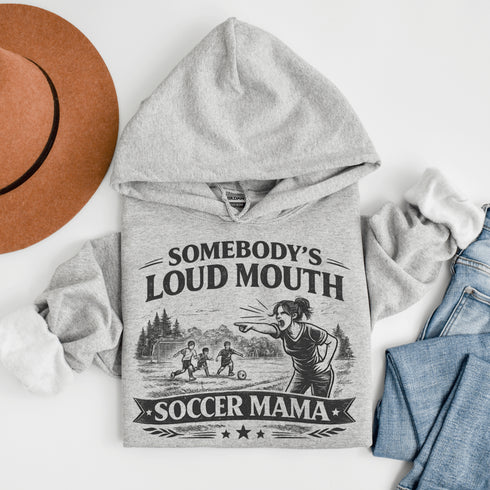 woman wearing grey hoodie with “Somebody’s Loud Mouth Soccer Mama” design featuring mom yelling on soccer sideline with kids playing in background