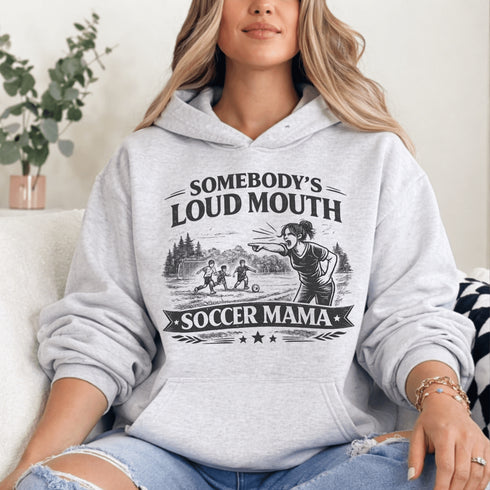 woman wearing grey hoodie with “Somebody’s Loud Mouth Soccer Mama” design featuring mom yelling on soccer sideline with kids playing in background