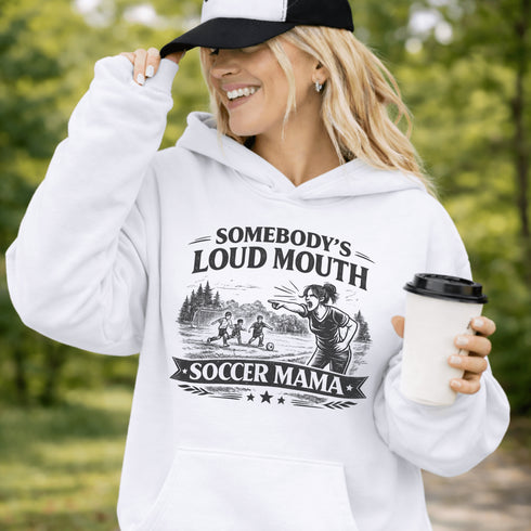 woman wearing grey hoodie with “Somebody’s Loud Mouth Soccer Mama” design featuring mom yelling on soccer sideline with kids playing in background