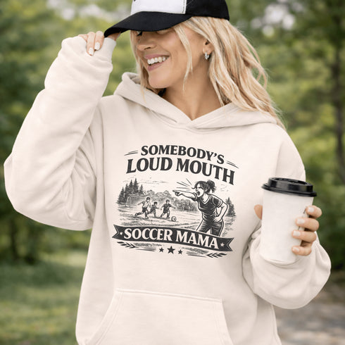 woman wearing grey hoodie with “Somebody’s Loud Mouth Soccer Mama” design featuring mom yelling on soccer sideline with kids playing in background