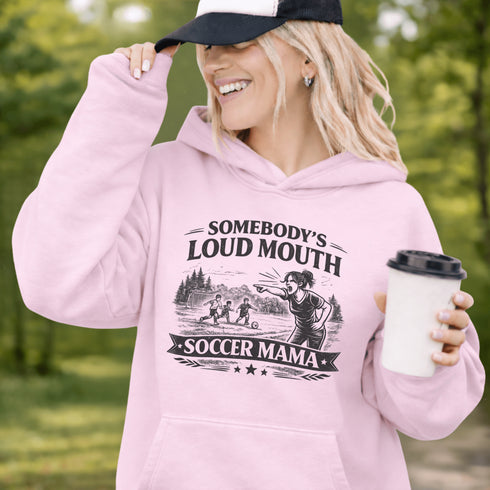 woman wearing grey hoodie with “Somebody’s Loud Mouth Soccer Mama” design featuring mom yelling on soccer sideline with kids playing in background
