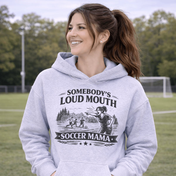woman wearing grey hoodie with “Somebody’s Loud Mouth Soccer Mama” design featuring mom yelling on soccer sideline with kids playing in background