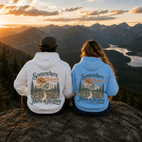 Two people sitting on a rocky mountain peak, seen from behind, wearing matching hoodies with a scenic back graphic that reads 'Somewhere Between Here & Heaven' over a mountain landscape. The person on the left wears a white hoodie, and the person on the right wears a light blue hoodie.