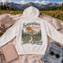 A white pullover hoodie laid flat on a pink blanket outdoors, with a mountain meadow and snow-capped mountains in the background. The hoodie has a large retro-style scenic graphic on the back reading βSomewhere Between Here & Heaven,β with an airplane, trees, and mountain illustration.