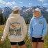Two women standing outdoors in a mountain meadow, wearing hoodies. The woman on the left is shown from the back, wearing a cream hoodie with a large graphic that reads 'Somewhere Between Here & Heaven' with a mountain and tree illustration.