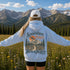 A woman stands in a mountain meadow with her back to the camera, wearing a light blue hoodie with a large graphic print on the back that reads 'Somewhere Between Here & Heaven' above an illustrated mountain, trees, and sunset. She is wearing a white cap and black shorts, with a backdrop of rugged mountains and pine forests.