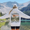 A woman stands with her back to the camera in front of a mountain lake, wearing a white hoodie with a large scenic back graphic that reads 'Somewhere Between Lost & Free' and a baseball player on a mountain. She is also wearing a white cap and black shorts.