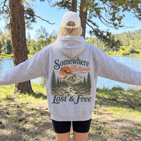 A person standing outdoors by a lake with trees and grass around them, wearing a light heather gray hoodie from the back. The hoodie has a large graphic print reading “Somewhere Between Lost & Free” with a mountain, pine trees, and an orange sun.