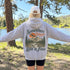 A person standing outdoors by a lake with trees and grass around them, wearing a light heather gray hoodie from the back. The hoodie has a large graphic print reading “Somewhere Between Lost & Free” with a mountain, pine trees, and an orange sun.