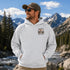 A man wearing a light gray heather hoodie with a 'Somewhere Lost & Free' graphic on the left chest, standing outdoors in front of a mountain and river landscape.